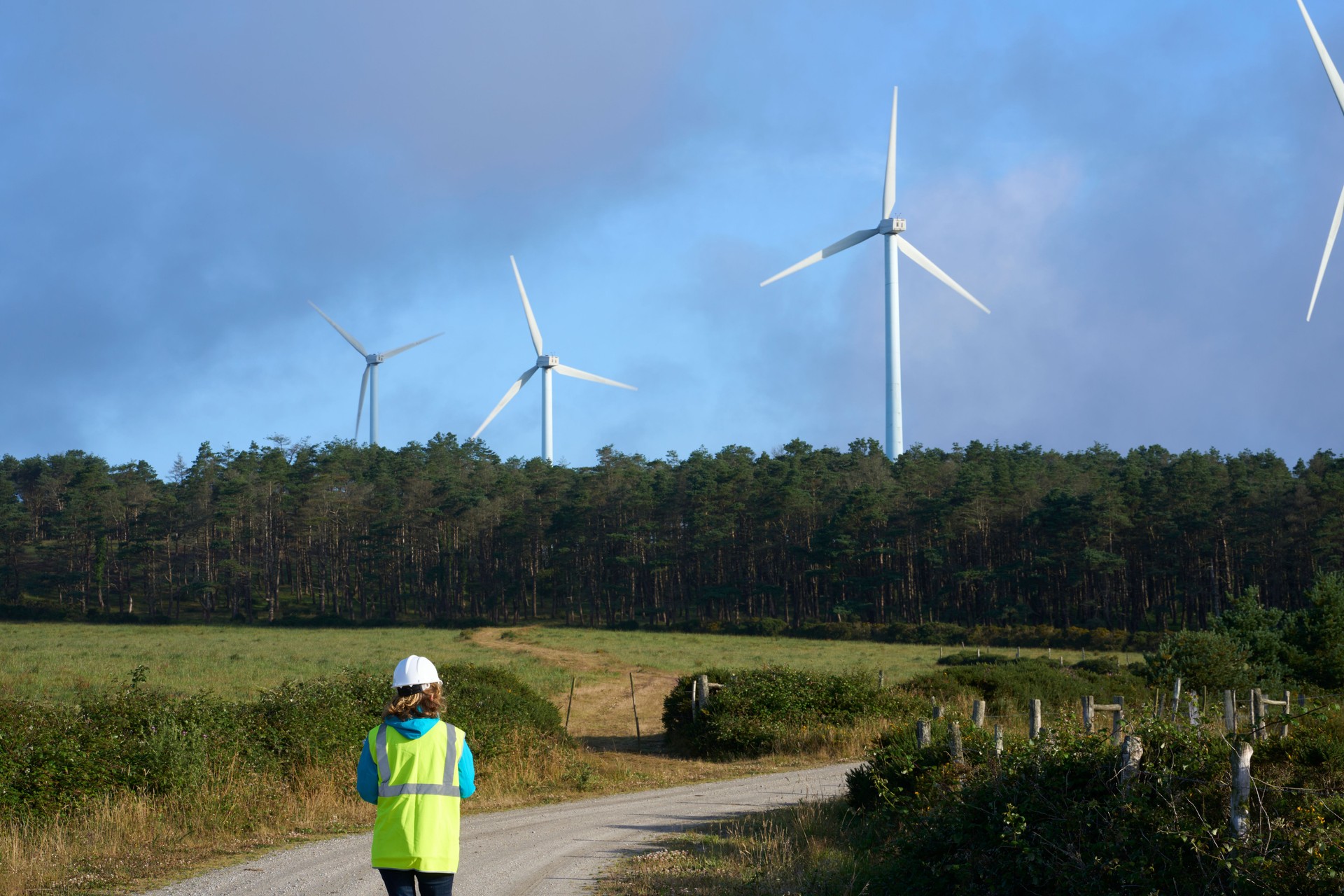 A worker with a white helmet and yellow vest goes to the wind turbines in a rural area.